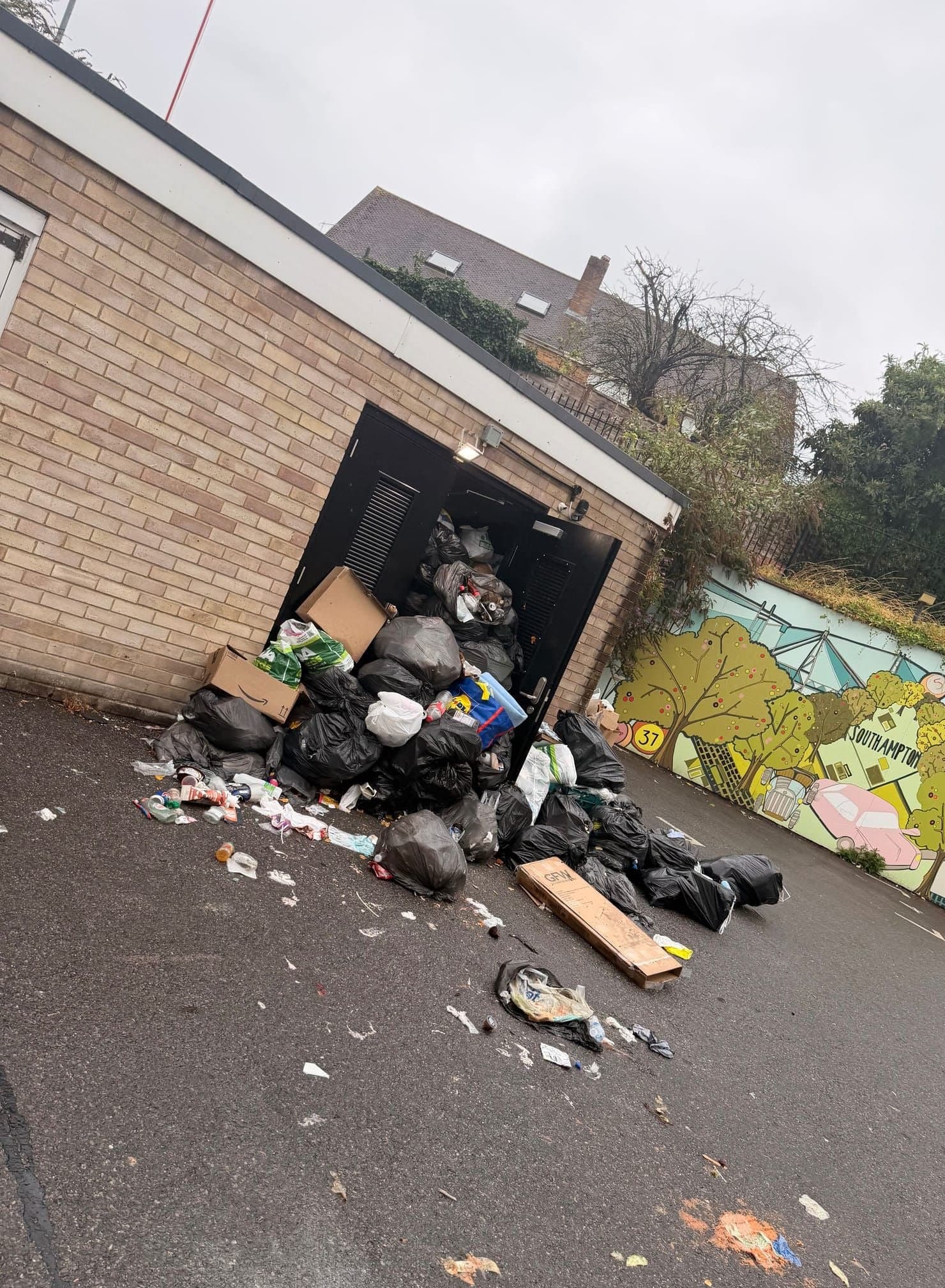 Bagged waste spilling out of a bin store doorway across a car park