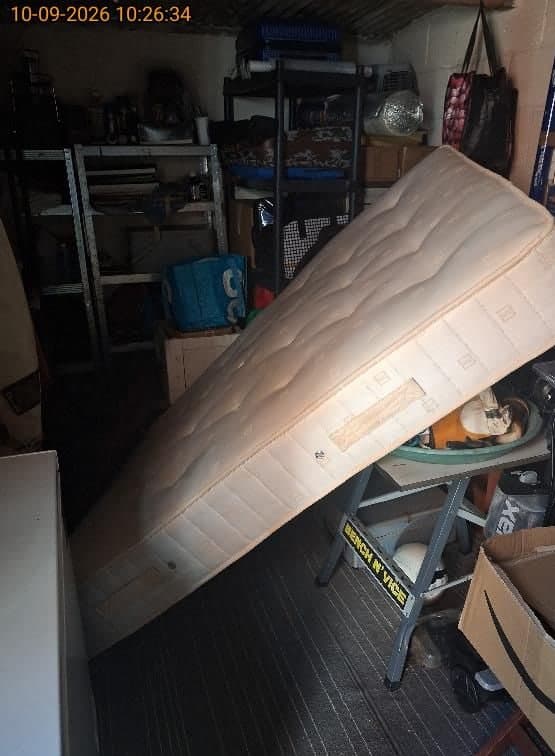 Old double mattress propped up beside cluttered shelving inside a garage awaiting clearance